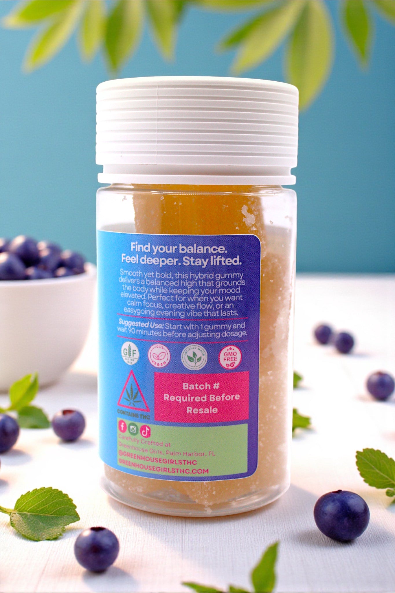 Jar of a product with blue label and white cap, surrounded by blueberries and green leaves on a light surface.