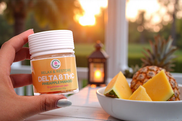 Hand holding a container labeled 'Delta 8 THC' with a bowl of pineapple and mango on a blurred outdoor background.