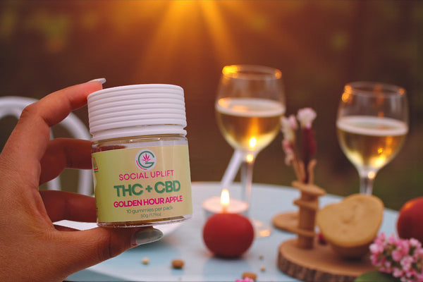 Hand holding a jar labeled 'THC + CBD' with two glasses of wine in the background.