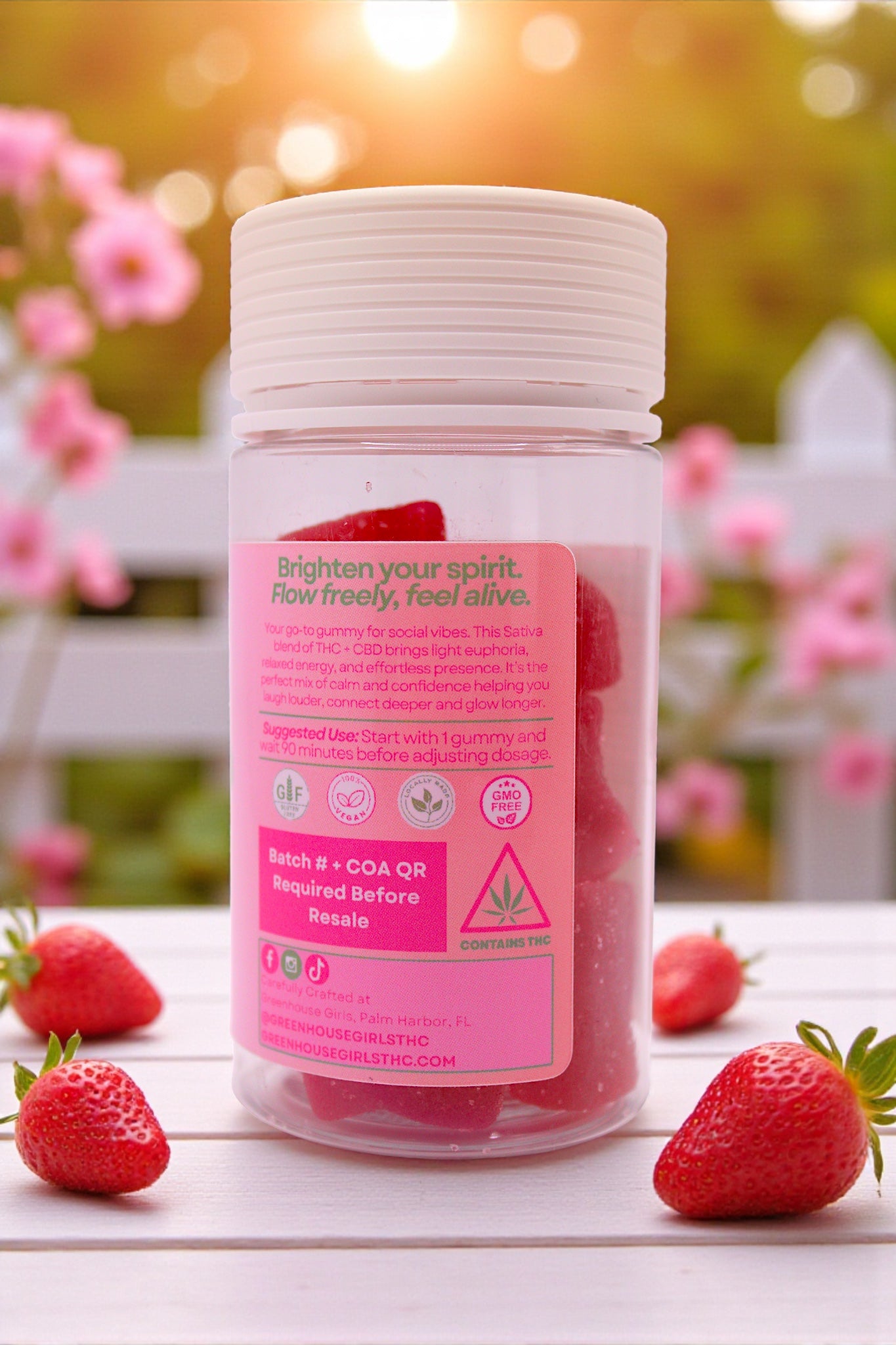 Bottle of supplements with a pink label surrounded by strawberries on a wooden surface with a blurred floral background.