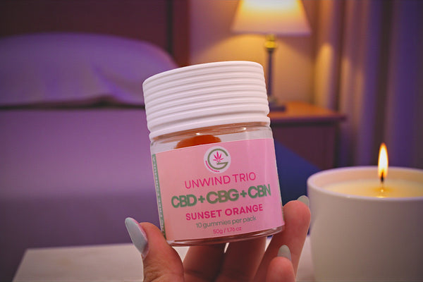 Hand holding a jar labeled 'Unwind Trio' with a pink label in a bedroom setting.