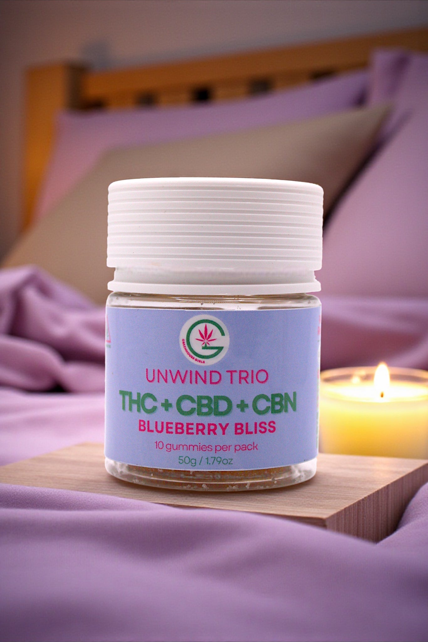 Jar of 'Unwind Trio' THC+CBD+CBN Blueberry Bliss on a wooden surface with a candle in the background.