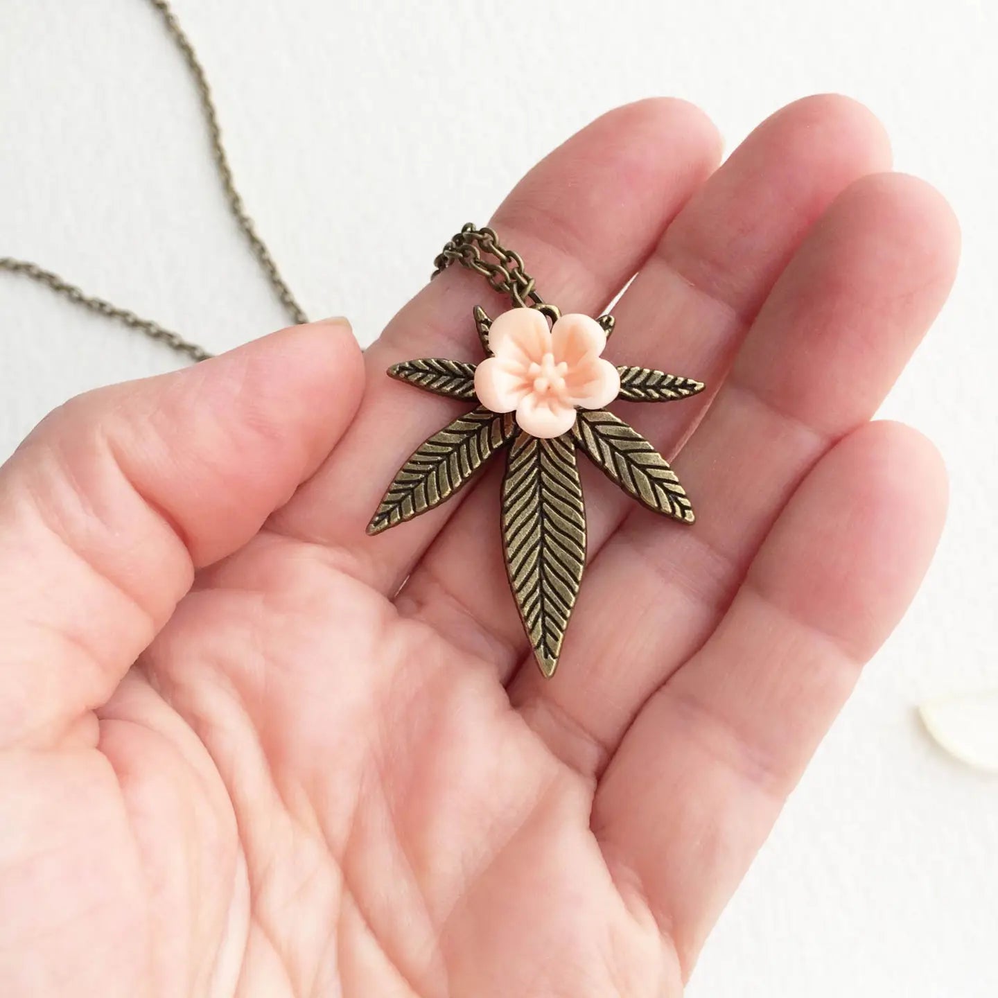 Hand holding a necklace with a leaf and flower pendant against a white background