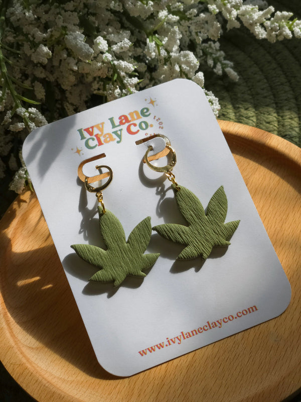 Greenhouse Girls handmade gold-plated huggie hoop earrings with green polymer clay cannabis leaf charms on display.